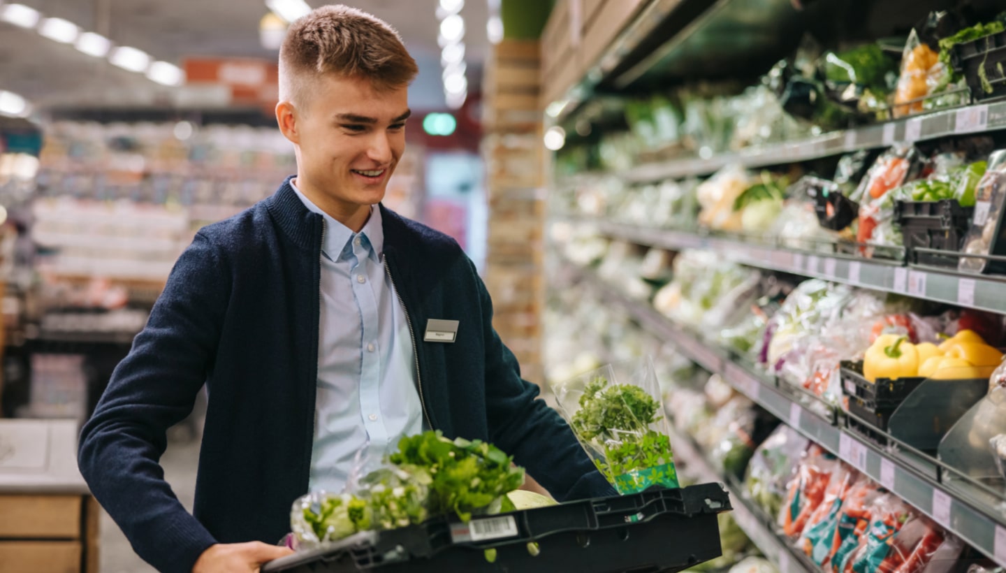 Ein Angestellte im Supermarkt räumt lächelnd Gemüse in ein Regal ein. Er hat gute Laune dank einer effizienten Dienstplangestaltung.