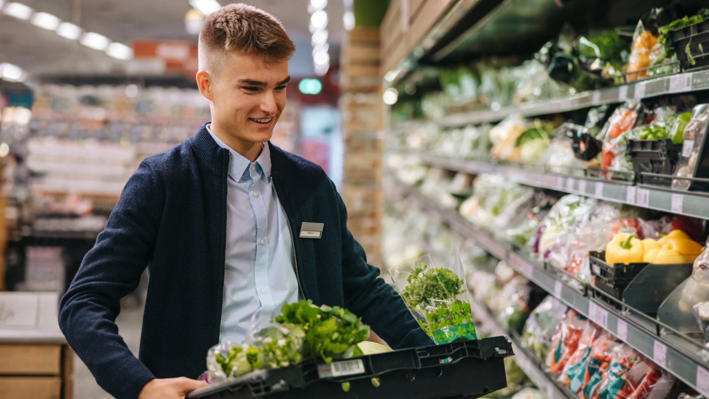 Ein lächelnder männlicher Supermarktmitarbeiter präsentiert stolz frisches Gemüse in einem Supermarktgang – ein Bild von Arbeitszufriedenheit und Stabilität inmitten der Herausforderungen der Mitarbeiterfluktuation."