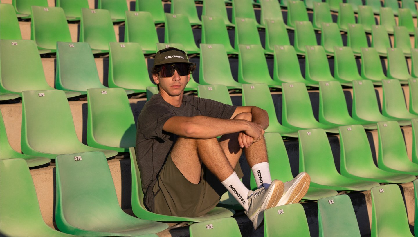 Männliches Model in grüner Salomon-Kleidung, mit Sonnenbrille und Cap, posiert auf einem Stuhl im Stadion. 